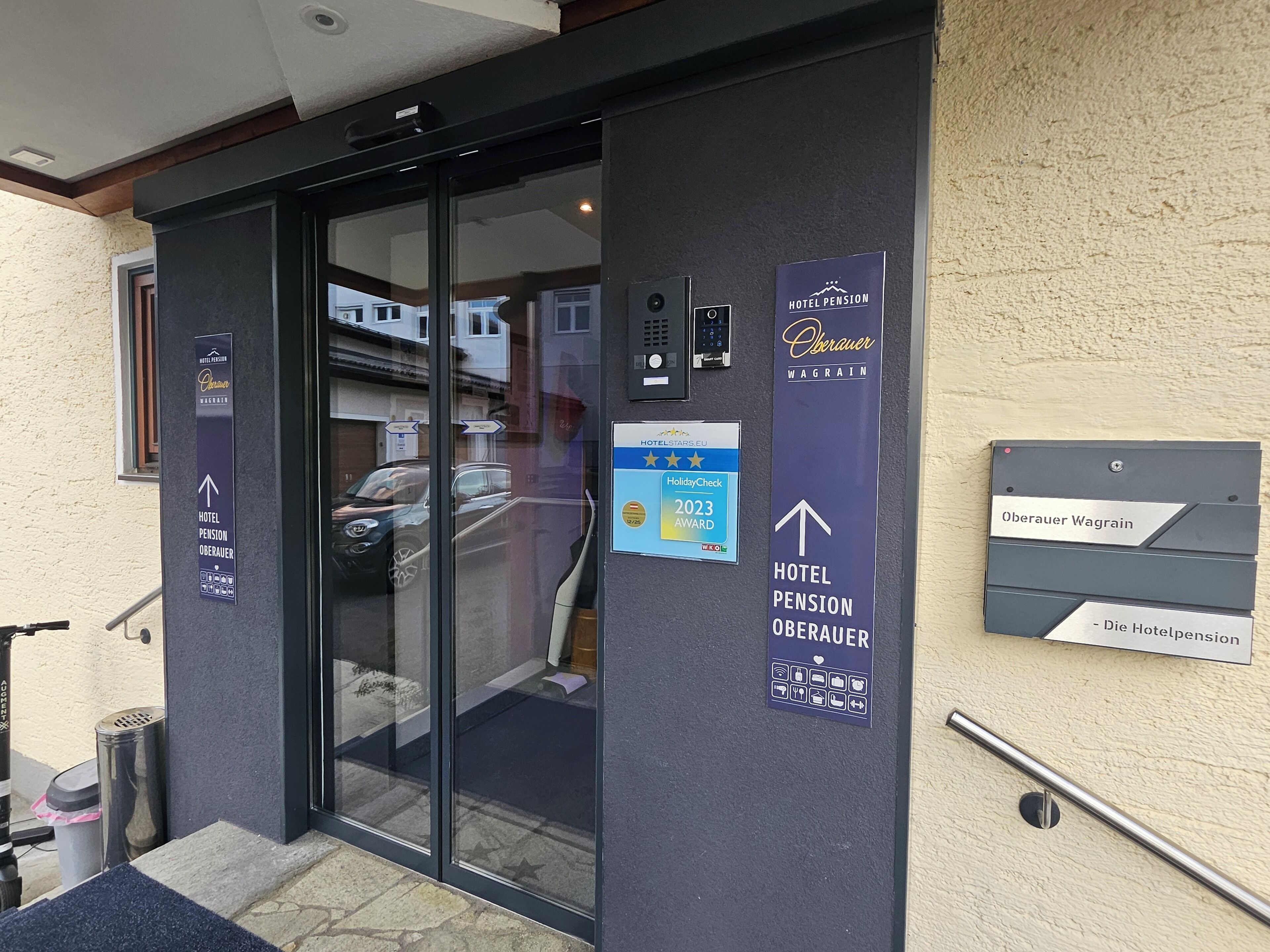 Een hotelentree met een glazen deur, borden die voorzieningen aangeven zoals een pension, en een plaquette voor het Oberauer Wagrain.