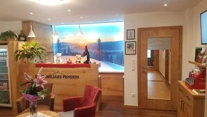 A reception area with a wooden desk, a red chair, a vase of flowers, and a view of a snowy landscape through a large window. - No1 - Flo's modern Rooms & Breakfast  (Wagrain)