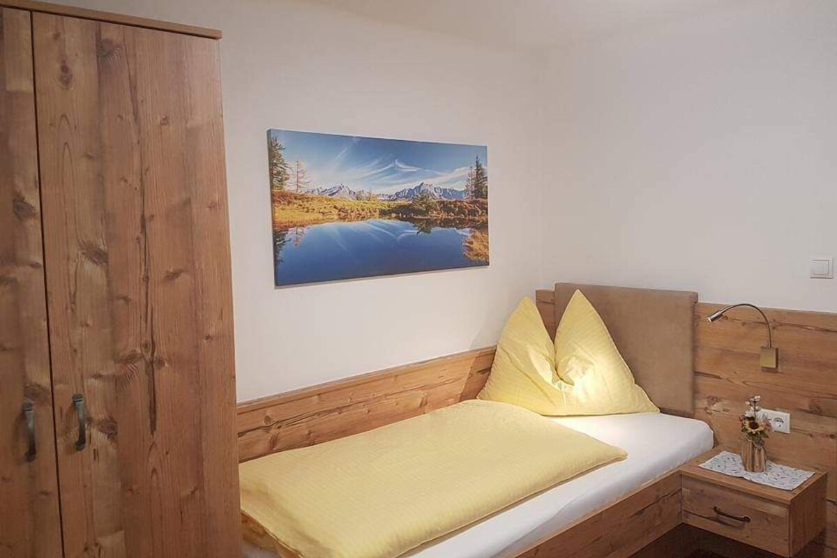 A bedroom with a wooden wardrobe, a bed with yellow bedding, a nightstand with a lamp, and a painting of a landscape on the wall.