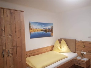 A bedroom with a wooden wardrobe, a bed with yellow bedding, a nightstand with a lamp, and a painting of a landscape on the wall. - No1 - Flo's Rooms & Breakfast  (Wagrain)