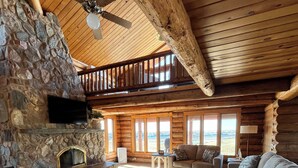 L’intérieur d’une cabane en rondins, avec une cheminée en pierre, un ventilateur de plafond et un coin salon confortable comprenant un canapé et des fauteuils.