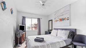 Ein Schlafzimmer mit Bett, Deckenventilator, Fenster mit Vorhängen, Flachbildfernseher und Wandbild.