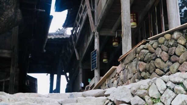 Un bâtiment traditionnel en bois, doté d’un mur en pierre et d’un escalier menant à un niveau supérieur.
