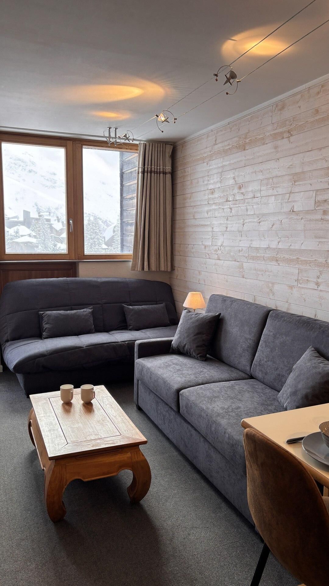 Una sala de estar moderna con un sofá gris, una mesa de centro de madera y vistas a un paisaje nevado a través de amplios ventanales.