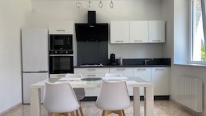 A modern kitchen with white cabinets, a black range hood, and a dining area with a white table and chairs. - Maison en Pierre (Ploumagoar)