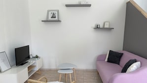 A modern living room with a flat-screen TV, a white sofa, a small round table, a wicker chair, and a pink couch. - Maison en Pierre (Ploumagoar)