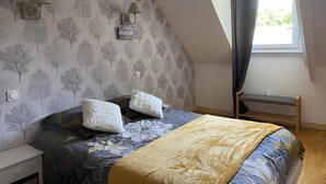 A bedroom with a sloped ceiling, a bed with a patterned duvet, two wall-mounted lamps, and a window with curtains. - Maison en Pierre (Ploumagoar)
