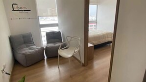 A modern, minimalistic living space with a grey sofa, a white chair, and a bed in the background. - Uhaus 704 (Manizales)