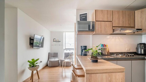 A modern kitchen with wooden cabinets, a microwave, and a coffee machine. There is a small island with a plant and a sign that says "Welcome." - Uhaus 704 (Manizales)