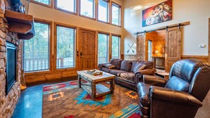 A spacious living room with a stone fireplace, large windows, a leather sofa, and a colorful rug. - Woodland Family Retreat Near the Lake (Broken Bow)
