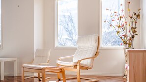 Une pièce avec deux fauteuils blancs rembourrés, un plancher en bois et un vase de fleurs placé près d’une fenêtre.