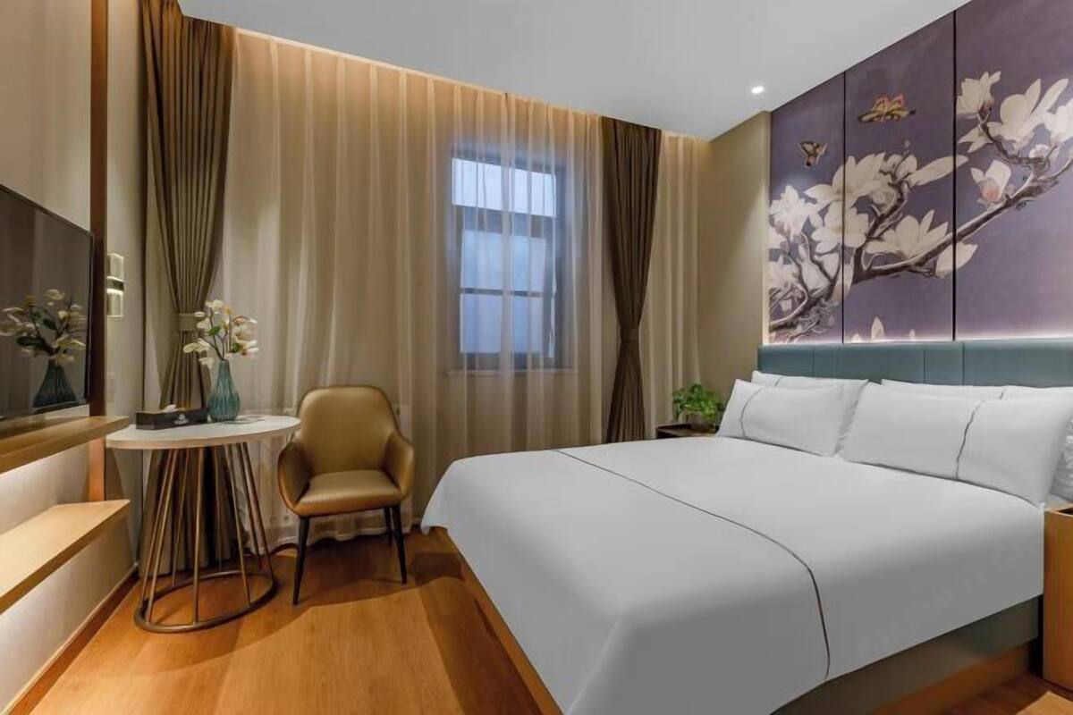 Une chambre d’hôtel comprenant un lit, un bureau, une chaise, une fenêtre avec des rideaux et un mur orné d’un tableau représentant des motifs floraux.