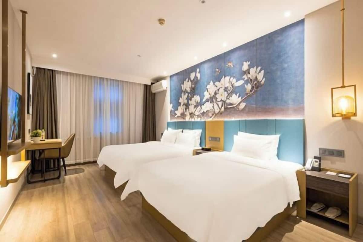 Une chambre d’hôtel avec deux lits, un bureau, une chaise, un tableau au mur et une table de chevet avec une lampe.