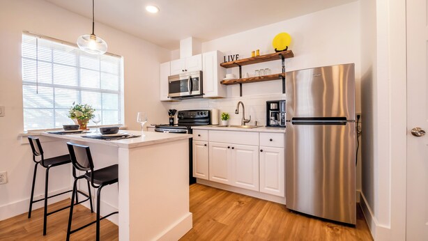 A modern kitchen with white cabinets, stainless steel appliances, a central island with bar stools, and a window with blinds.