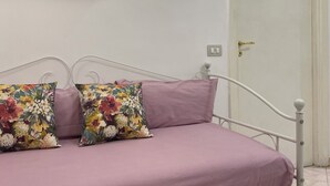 Een bed met een wit metalen frame, een paars matras en twee bloemenkussens. Er hangt een lamp boven het bed en bevindt zich een deur aan de rechterkant.