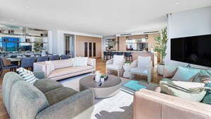 A modern living room with a sofa, armchairs, and a coffee table. There is a kitchen area in the background with a dining table and chairs.