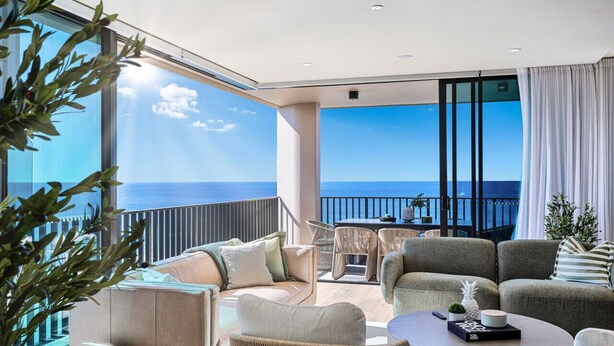 A modern living room with a large balcony overlooking the sea, furnished with sofas, a coffee table, and a dining area.