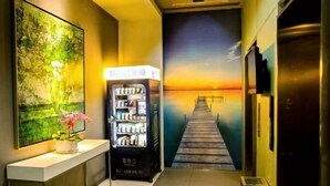 A hallway with a painting of a sunset and pier, a vending machine, and a potted plant. - Jinjiang Inn (Weihai Liugong Island Scenic Area Weigao Square Branch) (Weihai)
