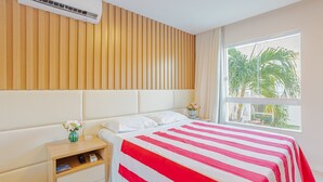 A hotel room with a bed, a nightstand, a vase with flowers, and a window with a view of palm trees. - GB105 Porto Smeralda Guarajuba a 150m da praia (Camaçari)