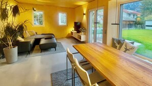 Una sala de estar moderna con una mesa de comedor de madera, un conjunto de sofás y una televisión. Hay dos ventanas que ofrecen luz natural y vistas al exterior.
