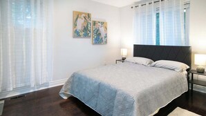 Une chambre moderne avec un grand lit, une table de chevet, deux œuvres d’art murales et une fenêtre ornée de rideaux légers.