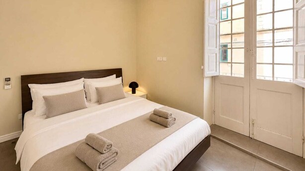 A hotel room with a bed, a nightstand, a lamp, and a window with a view of buildings. - Stylish 1 Bedr Apt next to The sea Front (Il-Gzira)