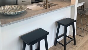 Une cuisine moderne équipée d’un réfrigérateur en acier inoxydable, d’une machine à café, d’un évier avec un robinet doré et de deux tabourets noirs.