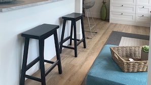 Une cuisine moderne avec des meubles blancs, un plan de travail en marbre et un téléviseur noir posé sur un meuble blanc. On y trouve deux tabourets noirs et un panier tressé sur un coussin bleu.
