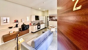 Una sala de estar moderna con un sofá gris, una pequeña mesa redonda y una zona de cocina con armarios blancos y un microondas.