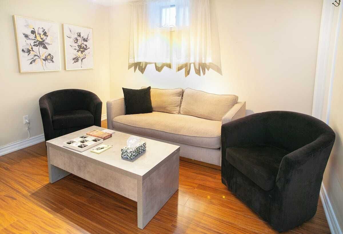 Een woonkamer met een beige bank, twee zwarte fauteuils, een salontafel met boeken en een decoratieve schaal, en twee bloemschilderijen aan de muur.