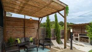 An outdoor patio with a wooden deck, a covered seating area with a sofa and chairs, and a pergola with hanging plants. - La Loc'Oyarn - Maison avec parking privé (Hennebont)
