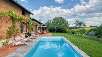 A swimming pool with lounge chairs, a brick building with vines, and a grassy area with trees.