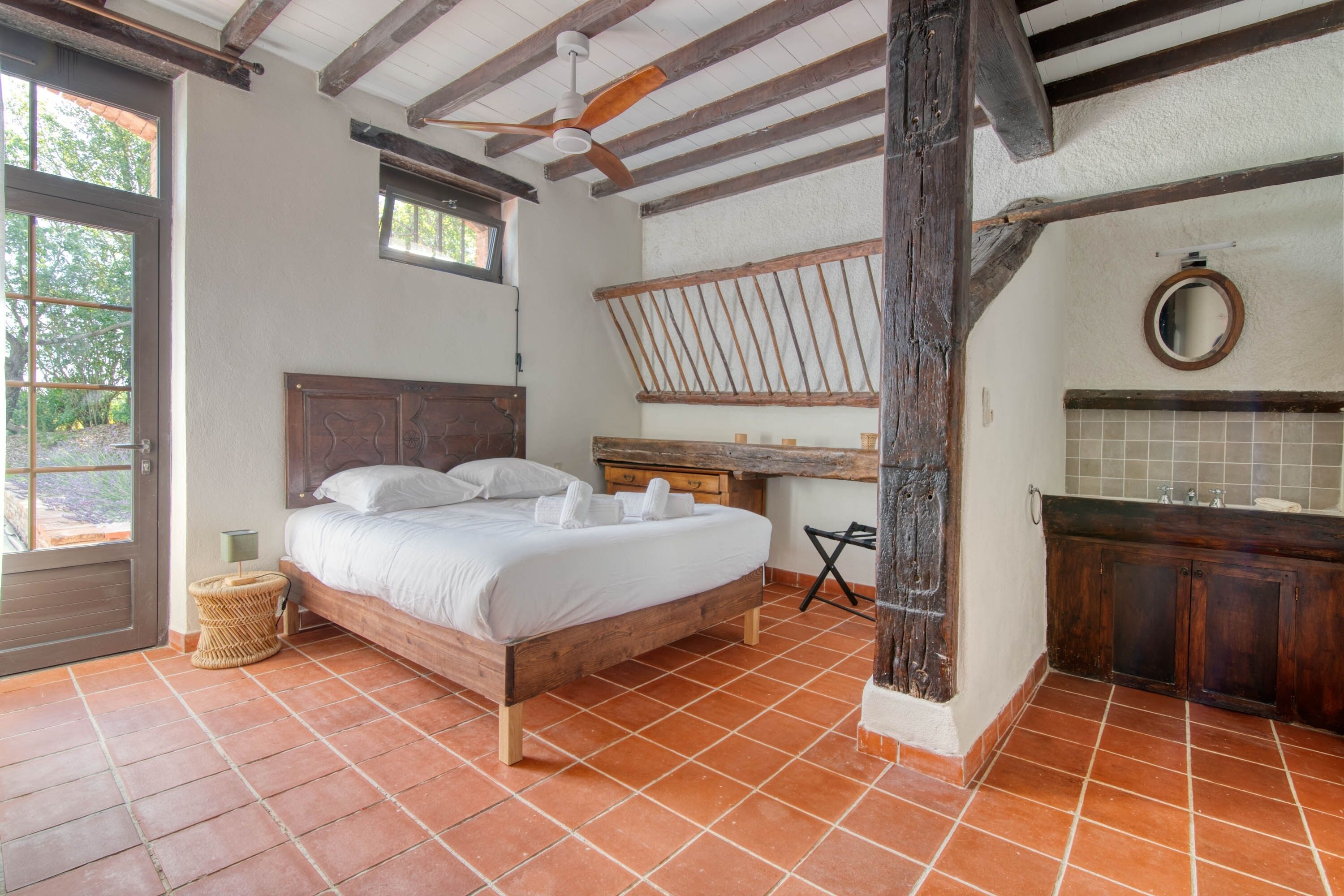 A bedroom with a wooden bed, a small desk, a round mirror, and a tiled bathroom.