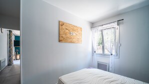 A bedroom with a bed, a window with curtains, a wall-mounted light switch, and a door leading to another room.