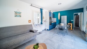 A modern living room with a grey sofa, a dining area with a glass table, and a kitchen with blue cabinets.
