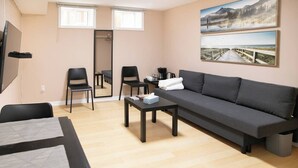 Una sala de estar moderna con un sofá, dos sillones, una mesa de centro y un televisor. Hay dos ventanas con persianas, un espejo y obras de arte en la pared.