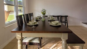 Una mesa de comedor dispuesta con cuencos y copas de vino, flanqueada por sillas y taburetes, con una ventana y vistas al follaje exterior.