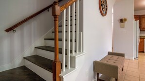 Una escalera con balaustres de madera y escalones oscuros, que conduce a un rellano con un banco acolchado y un reloj de pared.