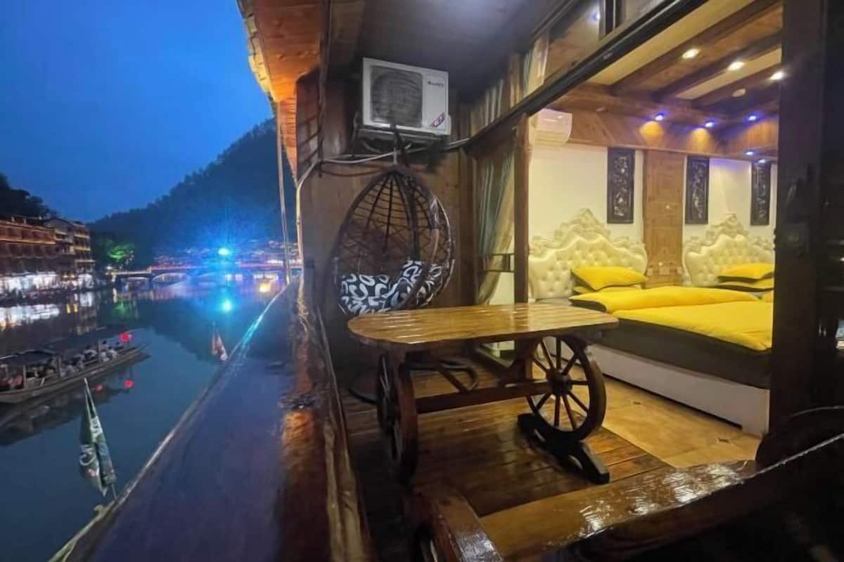 A wooden balcony with a table and chairs, a bed with yellow bedding, and a view of a river with boats.