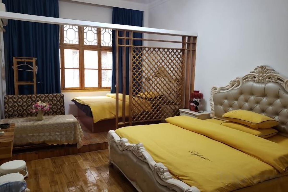 Una habitación con una cama grande, una cama más pequeña, un separador de madera, una ventana con cortinas y una mesita de noche con lámpara.