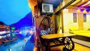 A wooden deck with a table and a chair, overlooking a river with boats and a mountainous backdrop.