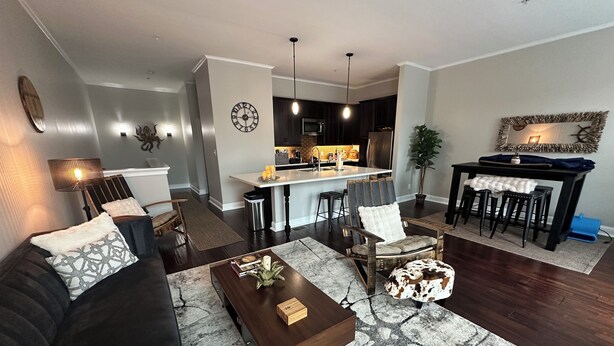 A modern living room with a dark wood floor, a black leather sofa, a marble coffee table, and a dining area with a wooden table and bar stools. - Charming 2bed, 2bath condo on The Strand, Galveston with WiFi, AC, garage (Galveston)