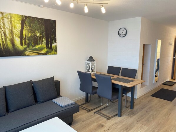 Ein modernes Wohnzimmer mit einer Couch, einem Esstisch, Stühlen, einer Uhr und einem Waldgemälde.