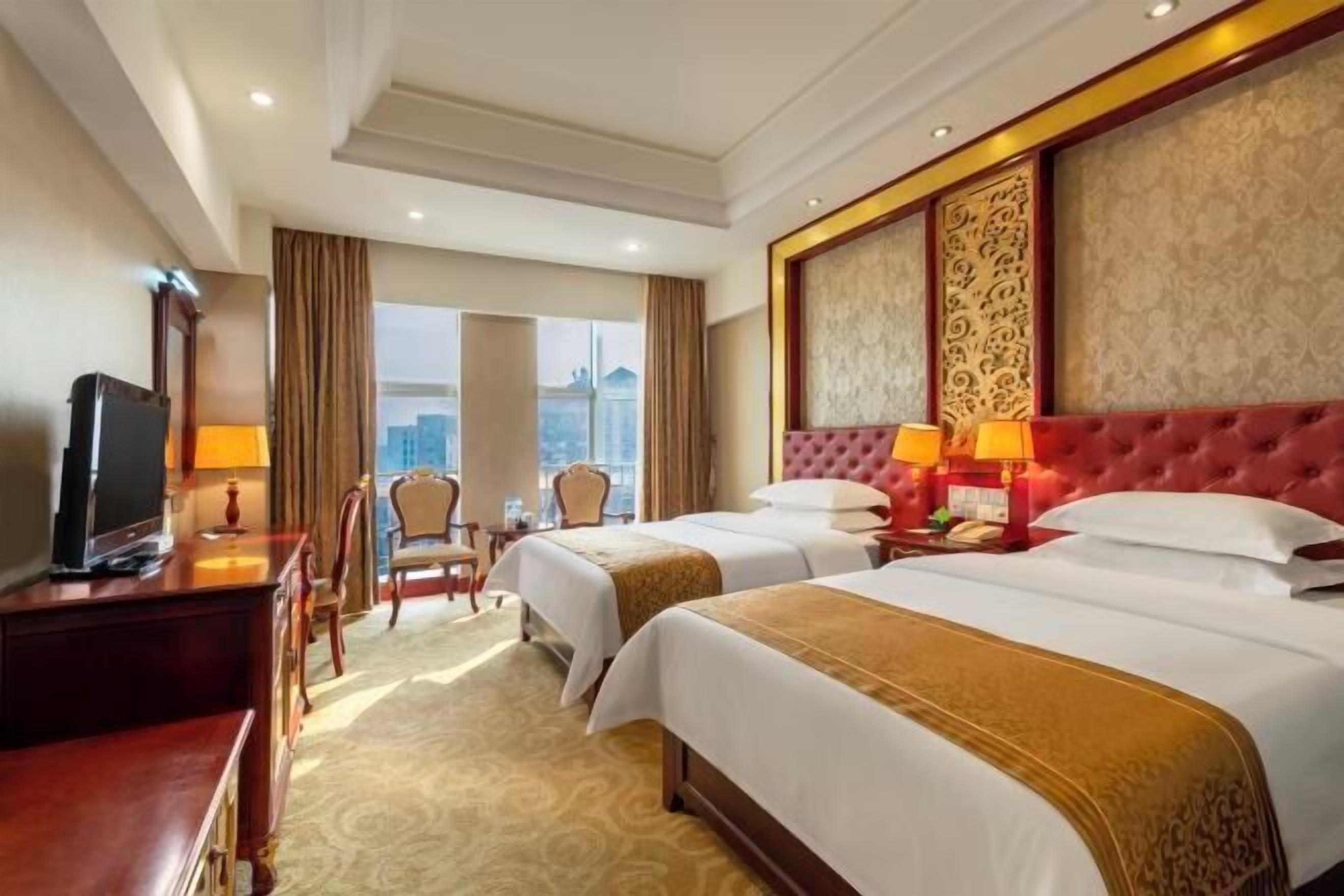 A hotel room with two beds, a television, a desk, and a view of the city.