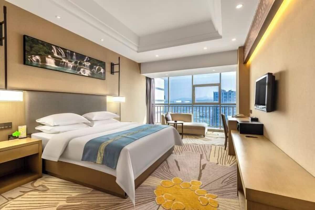 Une chambre d’hôtel équipée d’un lit, d’un bureau, d’une chaise, d’une télévision et d’une grande fenêtre donnant sur la ville.