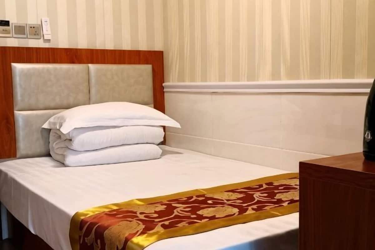 A neatly made bed with white linens and a red and gold patterned throw, a wooden headboard, and a wooden nightstand with a lamp.