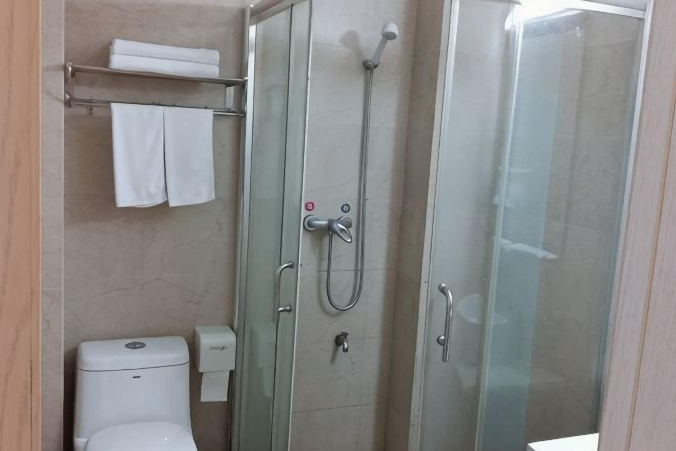 Cabina de ducha con paredes de vidrio, un cabezal de ducha manual, inodoro y un perchero con toallas.