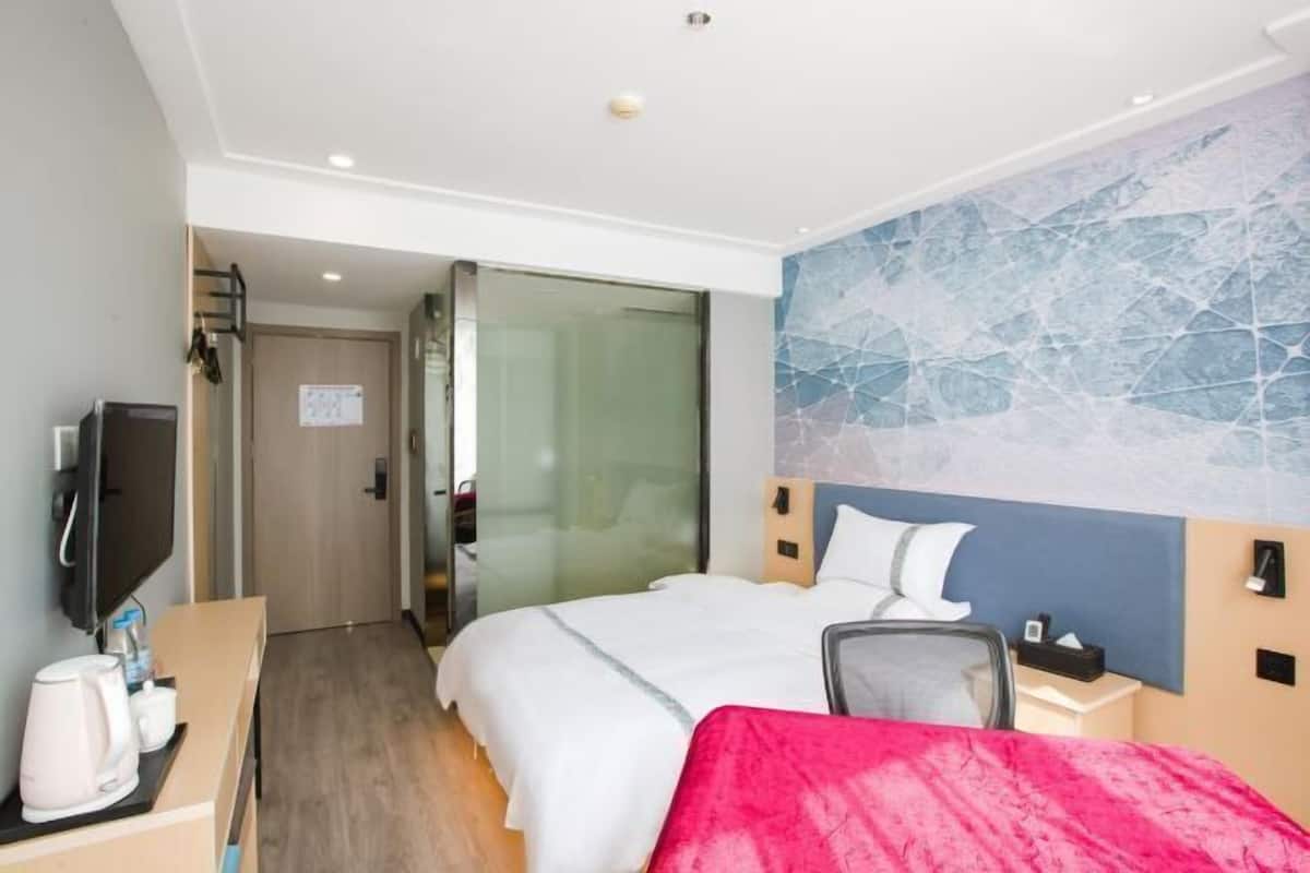 A hotel room with a bed, a desk with a coffee maker, a TV, and a glass door leading to a bathroom.