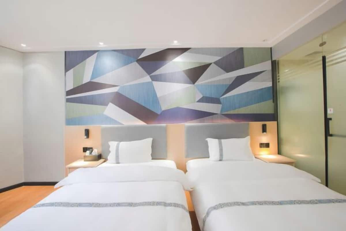 A hotel room with two beds, a geometric wall art, bedside lamps, and a glass partition.