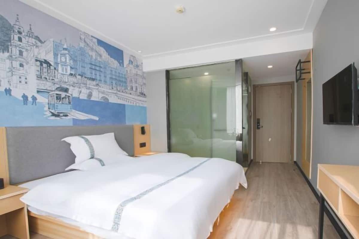 A hotel room with a bed, a painting of a cityscape, a television, and a sliding door leading to a bathroom.